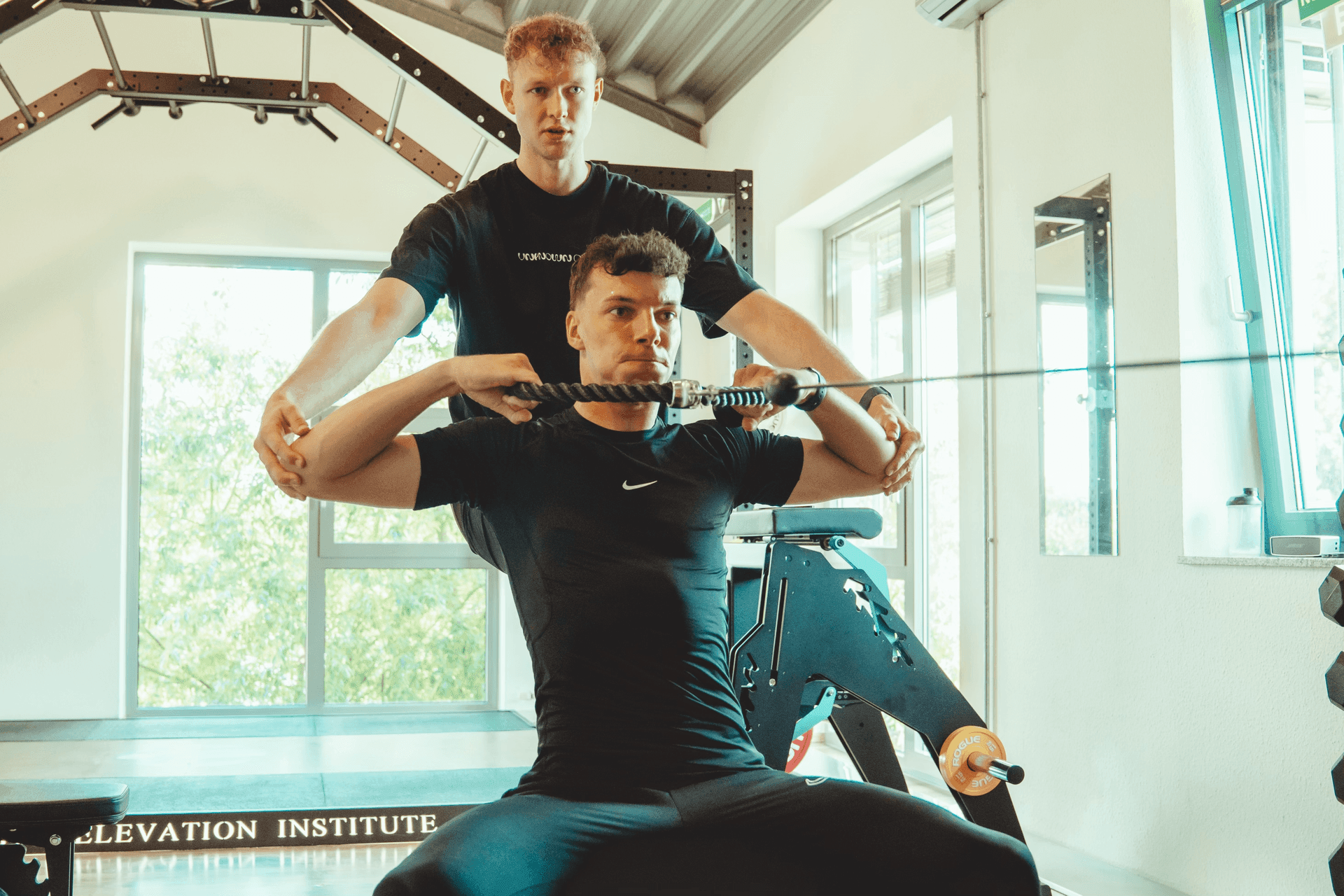 A trainer assists an individual using exercise equipment in a gym with large windows and natural light.